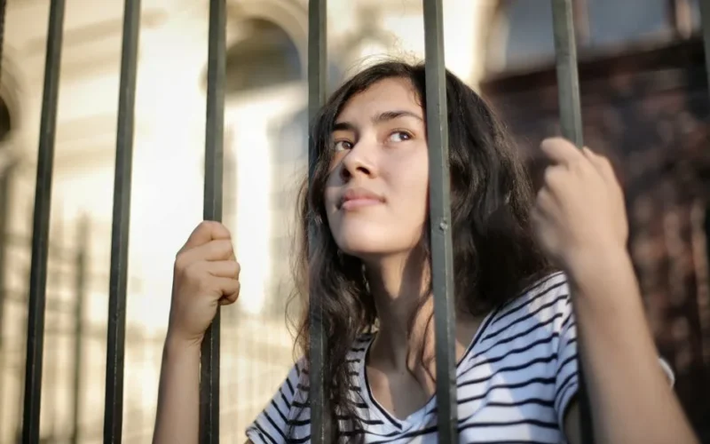 From below dreamy millennial lady in casual striped clothes looking up through iron bars of fence with hope as concepts of unhappy isolated extrovert and limited opportunities