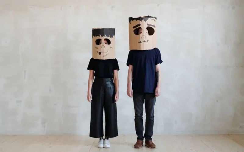 Couple wearing unique handmade cardboard masks as Halloween costumes in a modern setting.