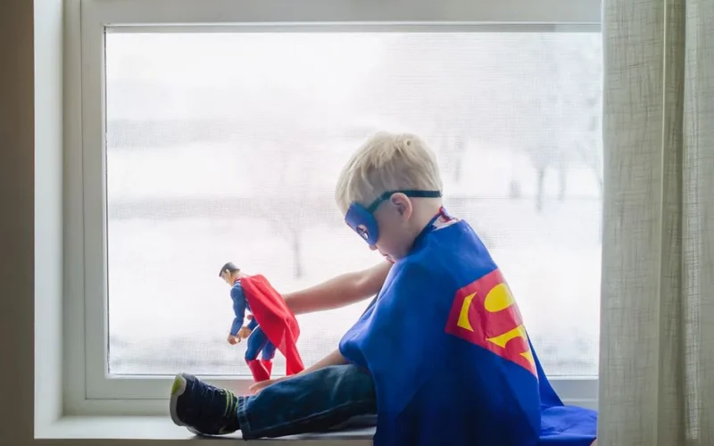 A young boy dressed as a superhero, playing with a toy by a window, embracing his imagination.