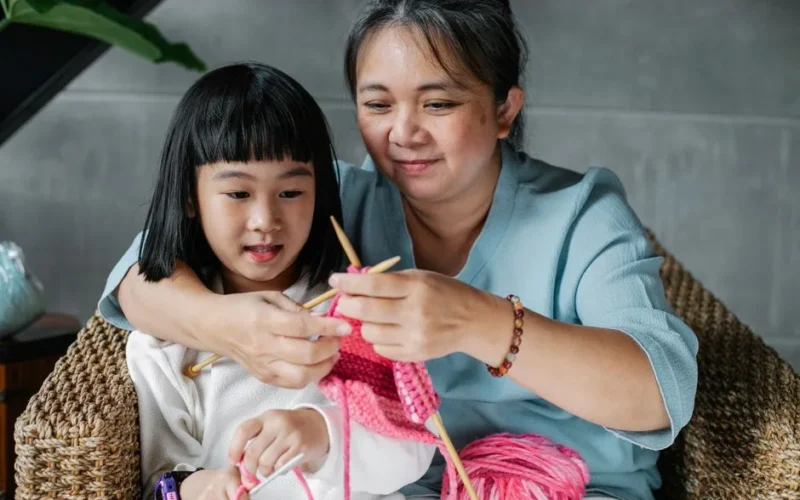 Content mature Asian woman embracing little granddaughter and explaining how to knit with needles
