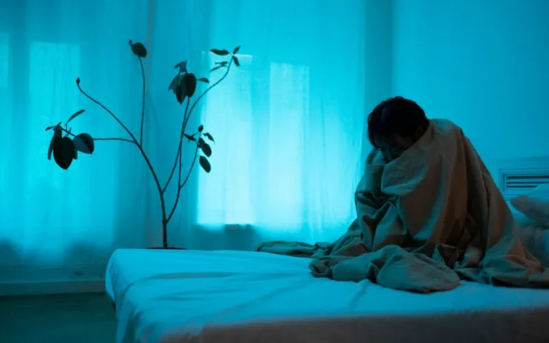 A woman sits alone on a bed wrapped in a blanket, conveying loneliness and depression.