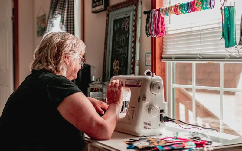 An adult woman sewing at home with a machine by a sunlit window.