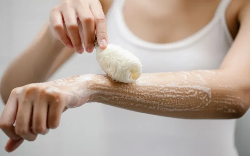 Unrecognizable female with shower sponge washing arm with foam while standing in light bathroom during daily skincare routine at home