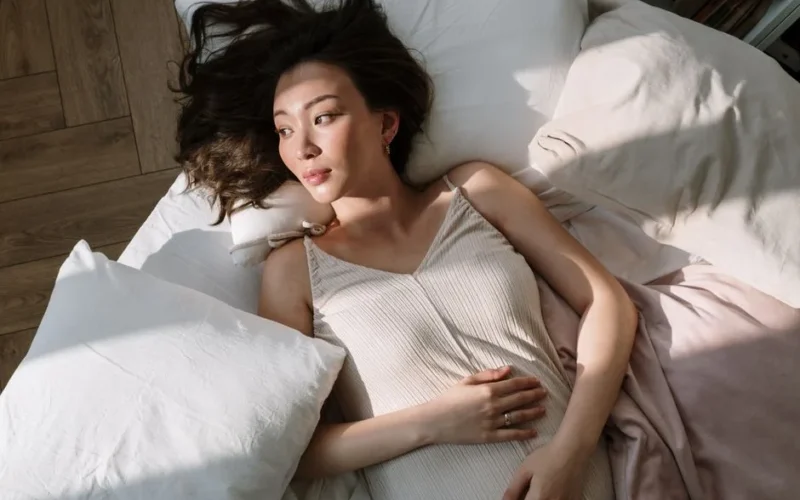 Asian woman in sleepwear lying on bed with hand on belly, enjoying morning sunlight.