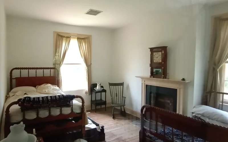 A small bedroom with 2 medium sized beds, and a fireplace. A small bedroom where visitors would sleep multiple people to a bed. Keywords: Mount Washington Tavern; National Road; Bedroom