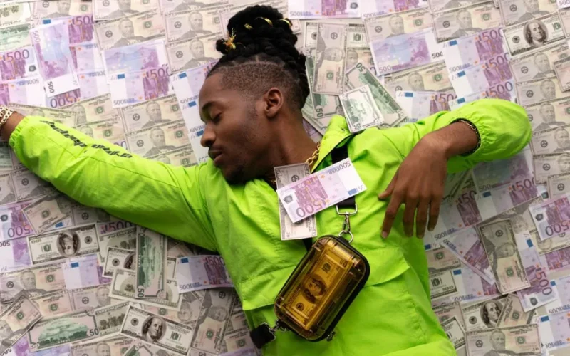 A stylish man in a green jacket lies on a pile of dollar and euro notes, symbolizing wealth.