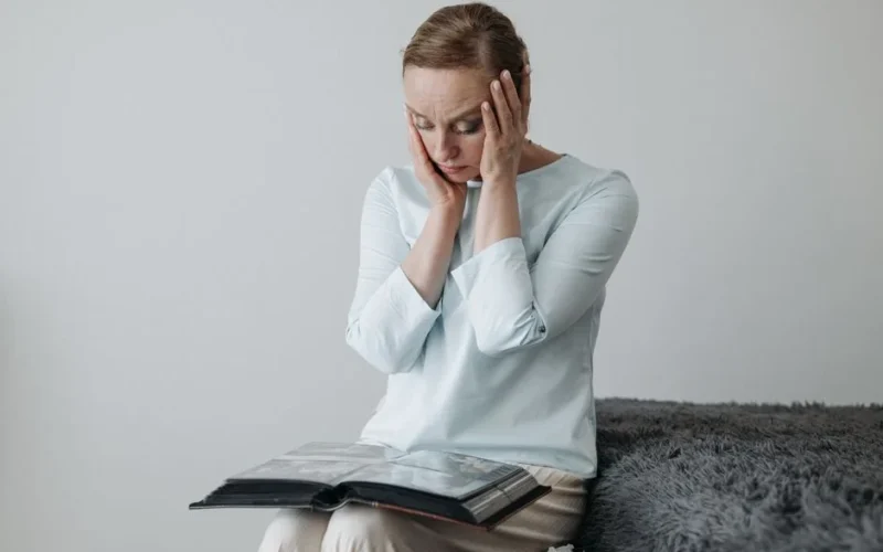 A woman sits thoughtfully with her hands on her face, looking at a photo album.