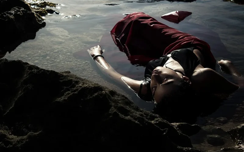 A young woman with a book on her face lies relaxing on a tree trunk by a tranquil lake.