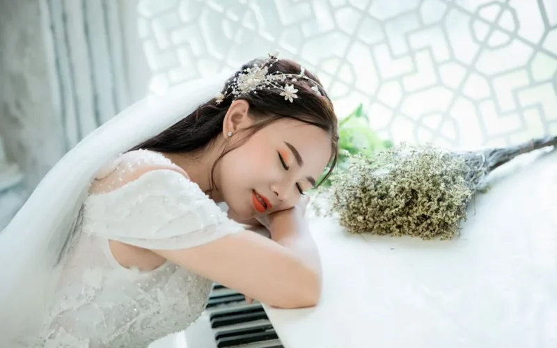 A serene bride in a white gown resting on a piano indoors, capturing elegance and tranquility.