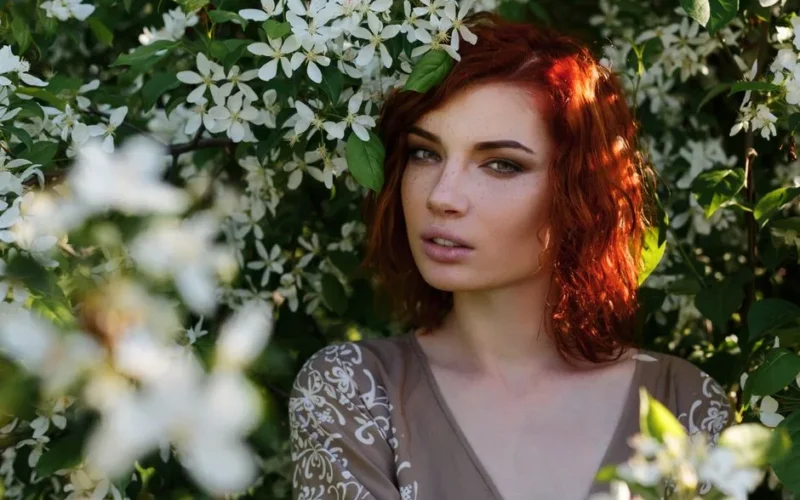 A redheaded woman poses among white flowers in sunlight, evoking a springtime feel.
