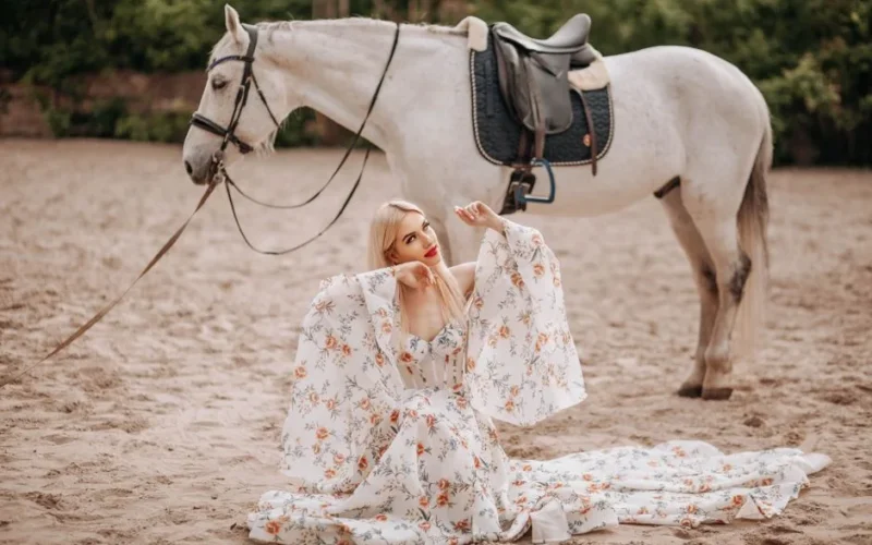 A woman in a floral dress sitting on sand next to a white horse, exuding elegance.