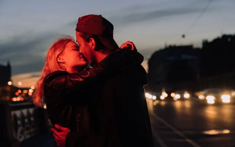 A couple sharing an intimate kiss on a city street during twilight, with blurred lights in the background.
