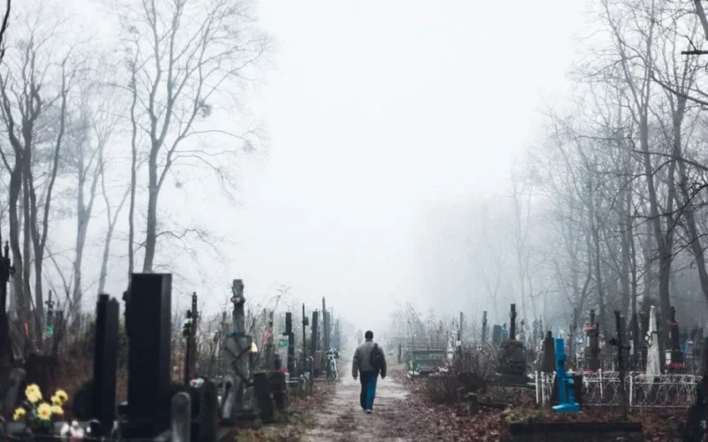 A lone man walks through a foggy cemetery surrounded by bare trees during fall.