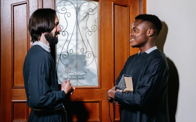 Two priests sharing a friendly discussion indoors, one holding a Bible, enhancing the spiritual atmosphere.