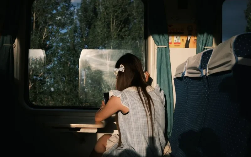 A young girl sits by the window of a train, enjoying the view outside. Sunlight casts a warm glow.