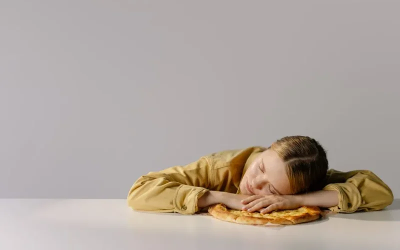 A Caucasian woman with eyes closed, resting her head on a pizza, embodying relaxation and humor.
