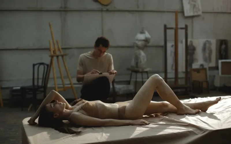 Artistic scene of a model posing nude in a studio with an artist sketching in the background.