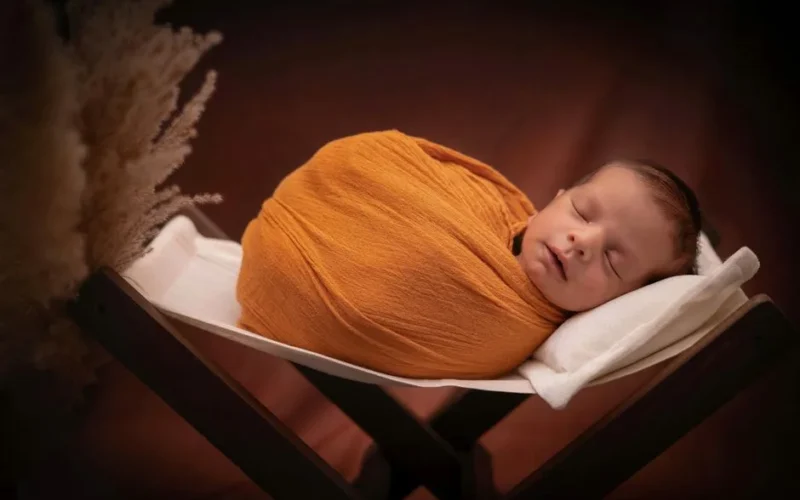 Adorable newborn baby peacefully sleeping in warm orange wrap on white cushioned basket.