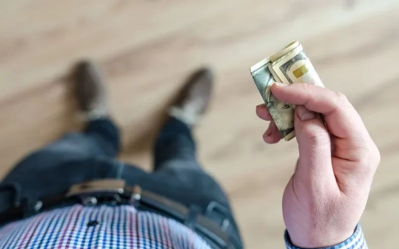 A person wearing denim and a checked shirt holds folded dollar bills in their hand indoors.