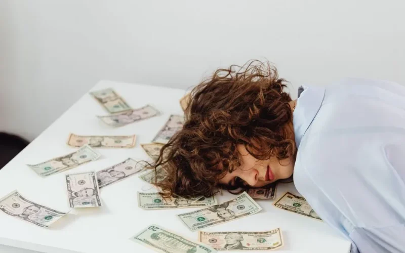 A woman with curly hair resting her head on a table covered in US dollar bills.
