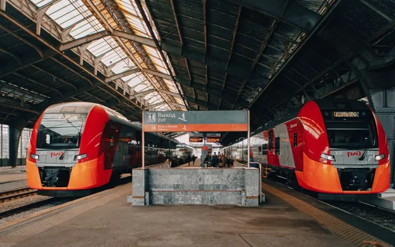 Red and black trains at a bustling railway station with passengers and exit sign.