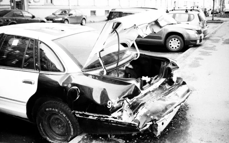 A black and white photo of a wrecked car on an urban street, highlighting vehicle damage.