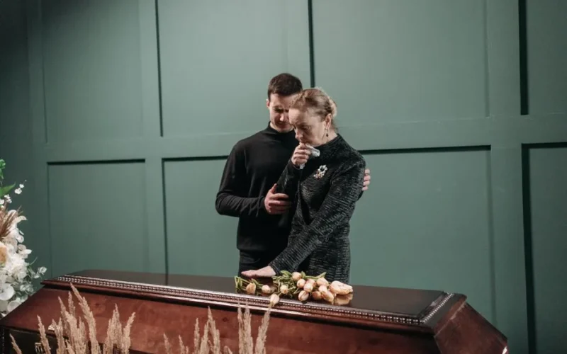 Grieving family members stand solemnly beside a coffin during a funeral.