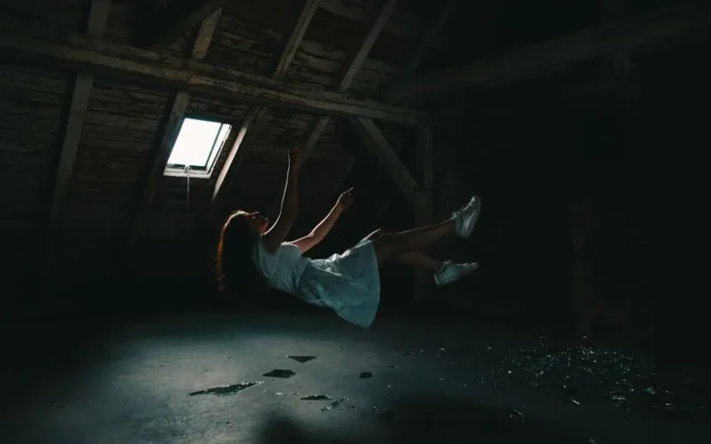 A surreal photograph of a woman in a white dress floating in an abandoned attic, capturing a sense of mystery.
