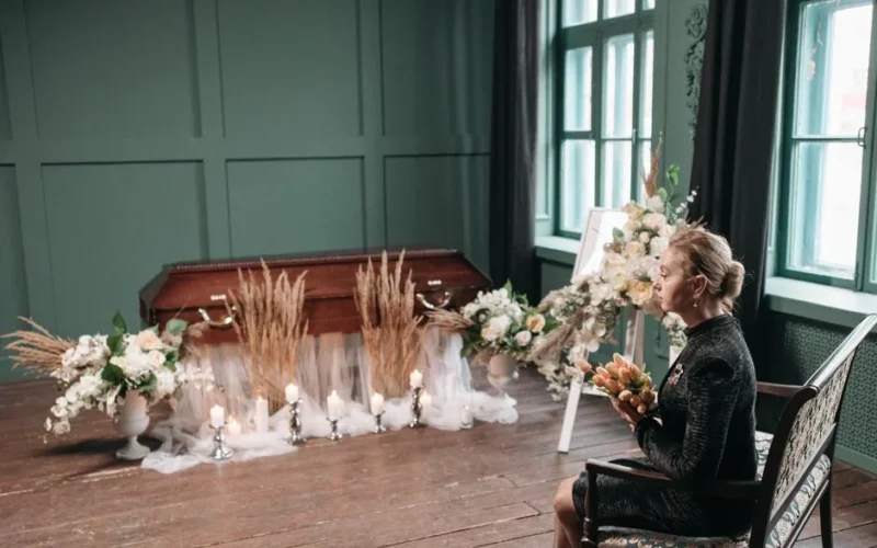 A woman in mourning sits near a coffin surrounded by flowers and candles at a funeral service.