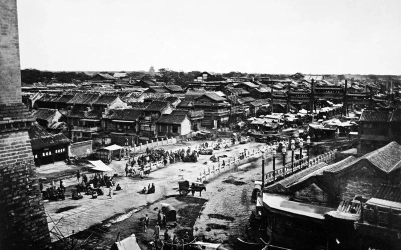 John Thomson: THIS view (No. 21) is taken from the city wall of Peking, close to the Ching-yang-men, or central gate, between the Chinese and Tartar quarters. It is in a direct line with the ' centre of the palace, and is the route which the Emperor traverses on his way to the Altar of Heaven. In the foreground we see a white marble bridge which spans a kind of city moat. This street, like all the thoroughfares in the Tartar city, is a very wide one, and is a place of great concourse and traffic. The lofty triple roof of the Temple of Heaven appears in the distance. The brick wall on the left is a portion of the great building which has its counterpart over every gate in Peking. These edifices were originally intended for storing ammunition, but the one shown here served for many years as a depository for the engraved wooden blocks employed in printing books. Formerly it contained the complete blocks for the collection of the Buddhist Scriptures, numbering over six thousand volumes, but these have been recently removed to a Buddhist temple in the north-east angle of the city. Wheeled vehicles arc forbidden to cross the centre of the bridge, this being reserved for the sole use of the Emperor. It is, however, a favourite resort for beggars — one among many such — and is known, therefore, to the European residents in Peking as the Beggars' Bridge. Here we may see these beggars gambling in groups, or stretched upon the pavement to expose their sores and nakedness to the public gaze. Many of these unfortunate and homeless beings are annually cut off by the keen frosts of early winter, and are found dead on the Imperial Bridge. There are a great number of stalls scattered along the principal streets, some of which are built like the old booths of the High Street of Edinburgh. In the centre of each street a raised causeway has been made, broad enough to accommodate two carts abreast, and intended for the carriage traffic. Between this raised causeway and the shops on either side of the road run broad spaces taken up with booths, tents, and stalls so closely packed as only to leave a narrow footpath close to the shops on the one side, and a chasm of deep mud pools on the other. From these pools material is taken for plastering and repairing the raised causeway. Most of these pools are stagnant and extremely polluted. One of my most disagreeable experiences during my visit to Peking was a ride along the road whilst the mud from these putrid pools was being ladled on to the highway to lay the dust. The dust, indeed, was laid, but fumes like those of the decomposing dead were raised in its stead. It would have been still worse had I, as some natives have done, lost my footing in a dark night, and been drowned in the mire. The verge of this slough teems with interesting life. Those who traffic in the stalls find many eager customers to buy their costly wares. I have seen one of these little booths, no better than that of some London costermonger, laden with over a thousand taels worth of jewels. The booths attract the attention of pedestrians. One finds in them all sorts of commodities for sale, and here and there are closely-packed crowds listening to the clever harangue of some auctioneer, who, with rare and ready wit, is extemporising rhyme in praise of cast-off silks, satins, or furs. In one I recollect a Mahometan butcher was plying his trade : the smiling follower of the prophet, encircled by carcases of sheep, ghastly heads, and entrails, stood behind a small wooden counter, knife in hand, giving a tender cut to a blooming slave girl, whose swarthy face was radiant with smiles, and whose cheeks were adorned with patches of rose-coloured paint. At another counter in this butcher's booth, a care-worn mother, with a nursing child in her arms, was fondling some miserable bones, and pleading with a greasy assistant for a bargain; but, frightful to behold, in the shade of the booth, my attention was riveted by a pair of gleaming blood-shot eyes, not indeed those of some ghastly head spitted on a meat-hook, but the hungry orbs of a street beggar, stark naked but for a thick coating of mud with which his body was smeared. A group of Peking street Arabs were tormenting this wretched being, until at last I saw him seize the ringleader, and daub him over with a ball of moist mud which he carried as a weapon. Adjoining this booth there was the tent of a dramatic reader, or story-teller, entertaining a well-dressed audience, seated before a long table on two rows of forms. At my approach he laid down his lute, and had a sly thrust at the loitering foreigner, which convulsed his hearers with laughter. Close by this was a cook's shed, with a series of brick ovens and fire-places in front. From these a powerful savour of roasting meats arose. Above a reeking caldron, puddings, spread on a clean board, were temptingly displayed. A group of boys and beggars were gathered in front to enjoy the pleasures of contemplation. The presiding genius of this cuisine, as he stuffed his puddings with their savoury contents, each time announced the fact in a shrill voice to the neighbourhood. His assistant was engaged with his left hand in kneading dough, while witli his right hand he twirled his rolling-pin on the board, so as to attract the notice of his customers. The agonized shriek of the master, and the twirling of the pin by the servant combined to maintain a constant stir and apparent bustle, which told upon purchasers. I presented a small boy with two cash. This gift enabled him at once to realize his fondest hopes. He invested in a reeking pudding, and, after a brief but affectionate look, devoured it with a relisii that was truly gratifying. The footpaths, close to the shops, exhibit a scene of great interest. One has frequently to follow along a narrow space left by a number of coal-laden camels, whose drivers are refreshing themselves at the nearest tea-shop, or else to make a dttottr to avoid damaging the wares which some shop-keeper has spread out upon the ground. Occasionally we brush past the cloth-covered sedan of some high mandarin, whose bearers and followers are shouting that the way must be cleared for the approach of The Great. After this, perhaps, a Tartar lady is encountered, rich in her Jewels and her silken attire. Her face, we can see, is carefully enamelled, and her lower lip is finished with a ravishing spot of vermilion, and behind follow her slave women, who add their own charms to their lord's seraglio, and who bear their mistress's purchases and the ills of their lot with equally stolid indifference. The liveried servants, or Yamen runners, hurry past in their conical extinguisher hats and red feathers, wearing in their faces an expression of sneaking contempt for the foreigner. Handicraftsmen ply their trades on every vacant space; and well-dressed merchants bustle to and fro, each intent on business of his own; whilst at every hundred steps at least an equal number of natives pass, who possess no distinctive characteristics, and of whom all that a foreigner can say is that they resemble their neighbours.