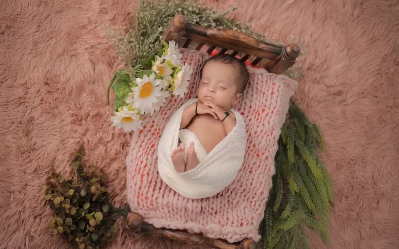 Adorable newborn baby wrapped in a blanket, peacefully sleeping on a wooden bed surrounded by flowers.