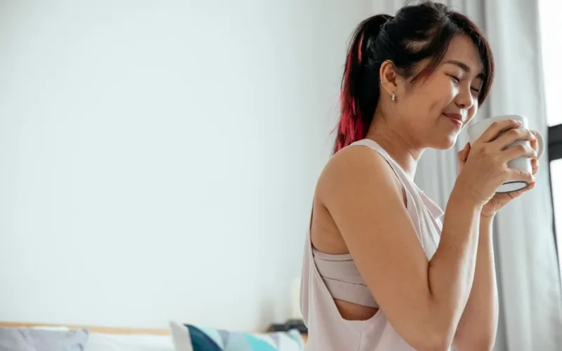 Side view of young Asian female in white shirt standing with closed eyes and enjoying aromatic coffee in morning at home