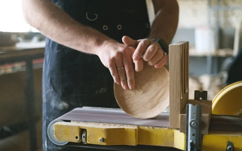 Unrecognizable male woodworker shaping round wooden detail while working at belt and disc sander in professional workshop on blurred background