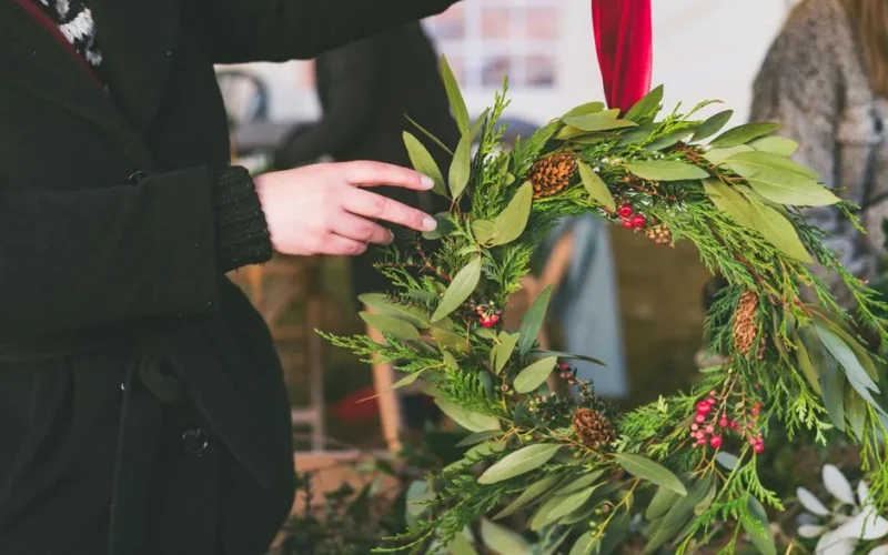 A woman decorates a holiday wreath with greenery, cones, and berries, adding a festive red ribbon.