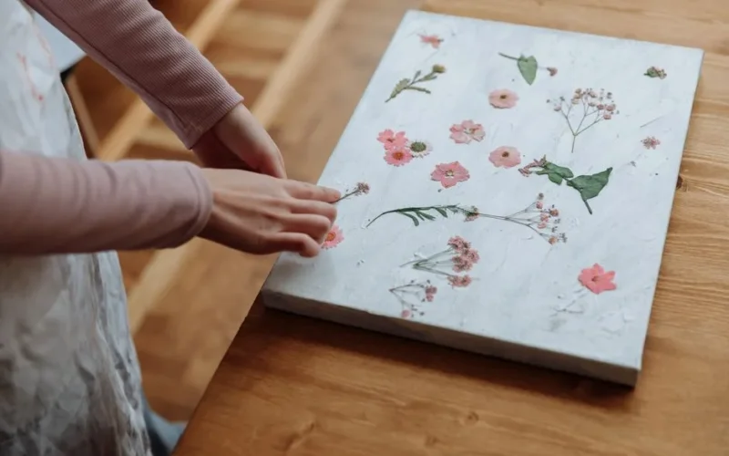 Hands arranging pressed flowers on a canvas indoors, highlighting a creative process.