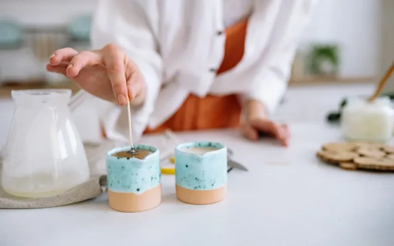 Close-up of hands making handmade candles using soy wax in decorative containers.