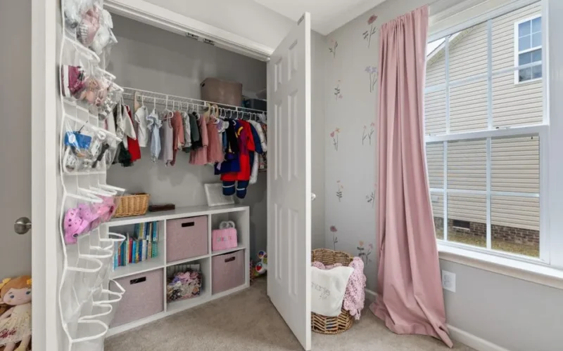 A neatly organized children's closet with clothes, toys, and pink decor in a bright room.