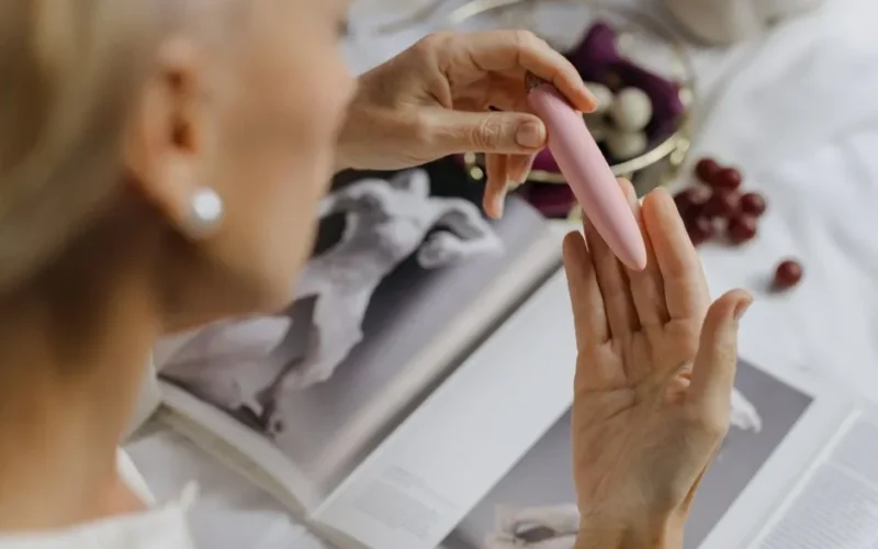 Close-up of a woman holding a pink vibrator, with art books in the background, emphasizing intimate and personal themes.