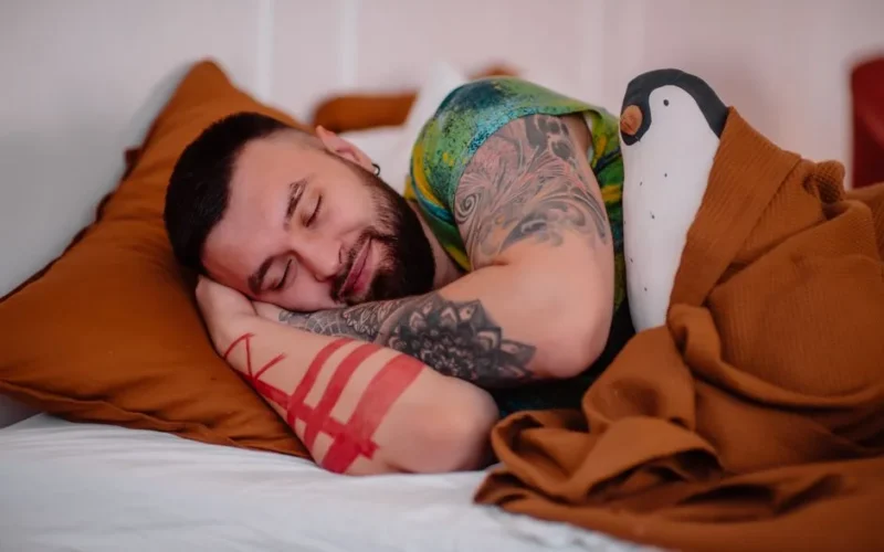 A tattooed man with closed eyes sleeps peacefully indoors, showcasing relaxation.