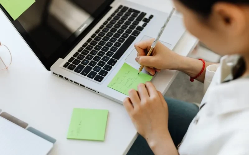Person writing a to-do list on sticky note next to laptop for organization.