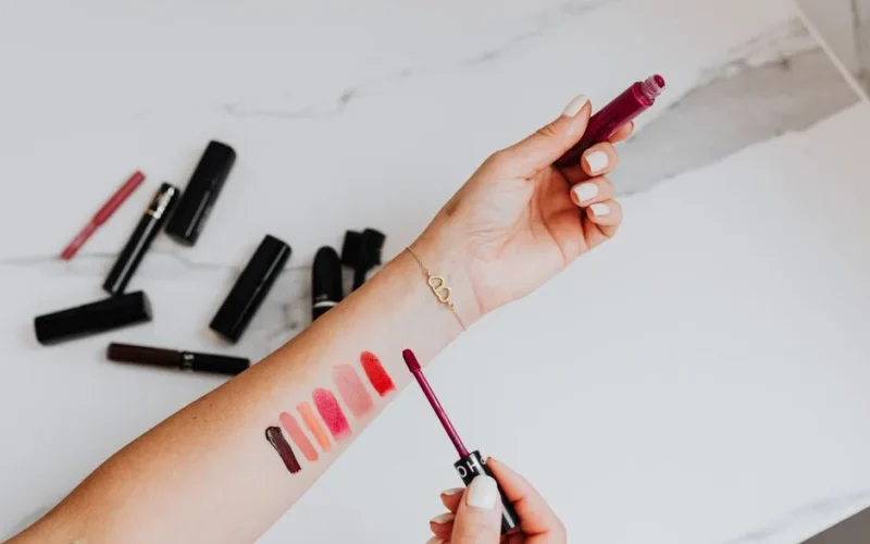 Woman applying and testing various lipstick shades on her arm over a marble surface.