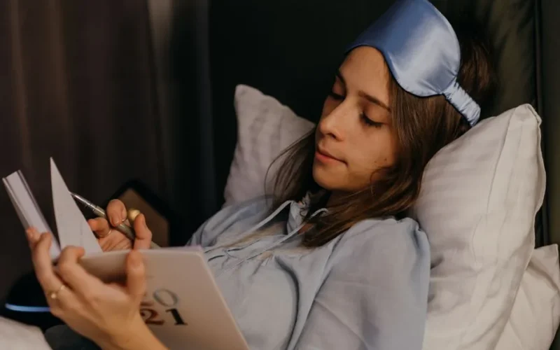 A young woman relaxing and writing in her journal while resting in bed with a sleep mask.