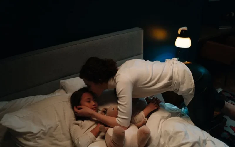 A mother tenderly kisses her sleeping child goodnight in a warmly lit bedroom, creating a peaceful scene.