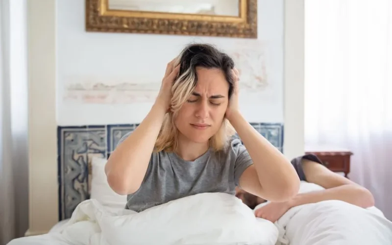 A woman holding her head in discomfort while sitting on a bed in a sunlit room.