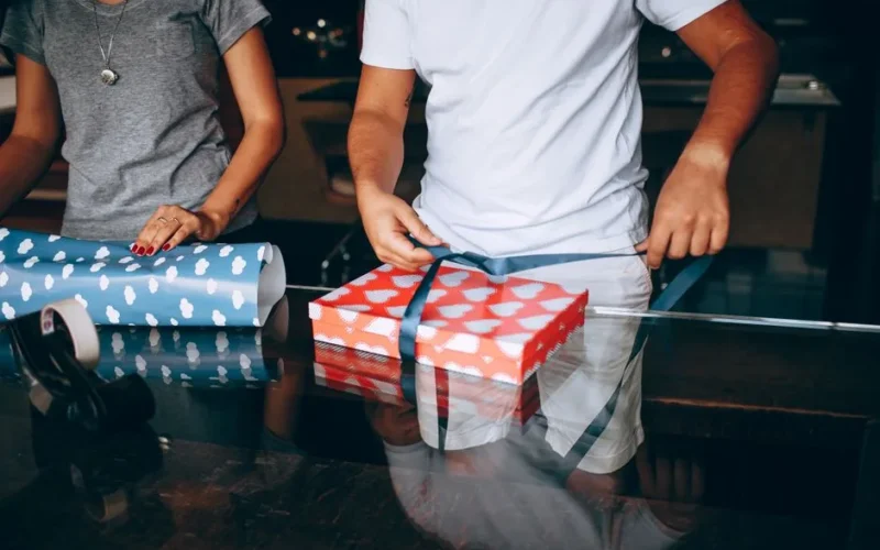 A man and woman wrapping a gift box with heart-patterned paper at home.