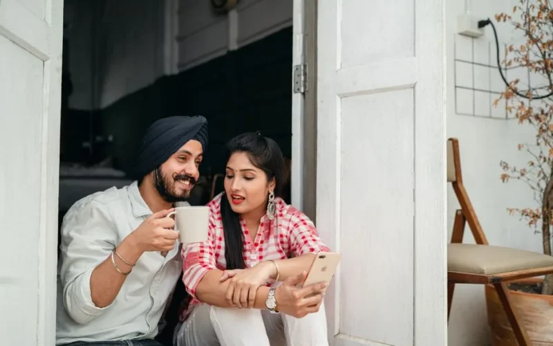 Cheerful young Indian man with coffee and elegant Indian lady with smartphone talking while sitting on floor on veranda
