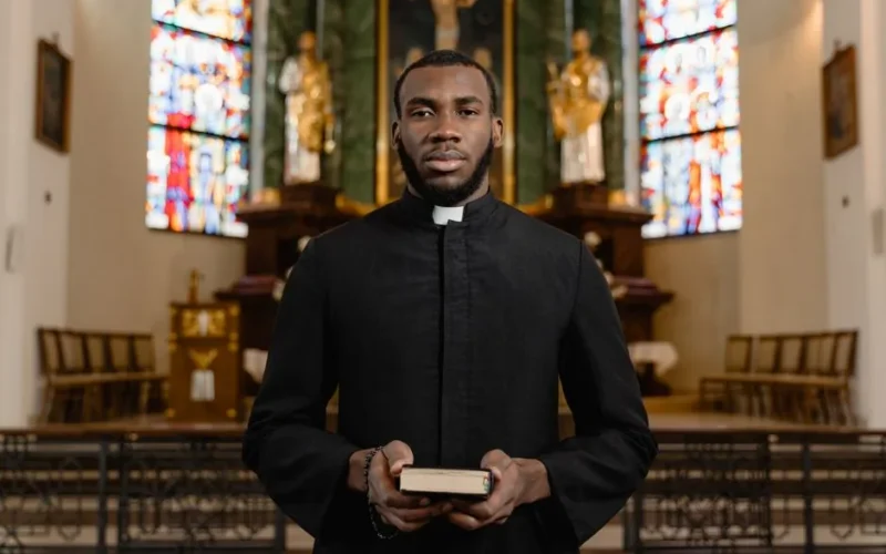 Portrait of a priest in a cassock holding a Bible in an ornate church interior.