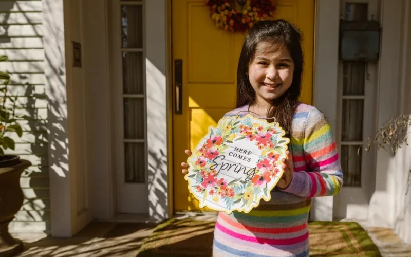 Smiling girl holding a vibrant spring decor sign at home entrance.