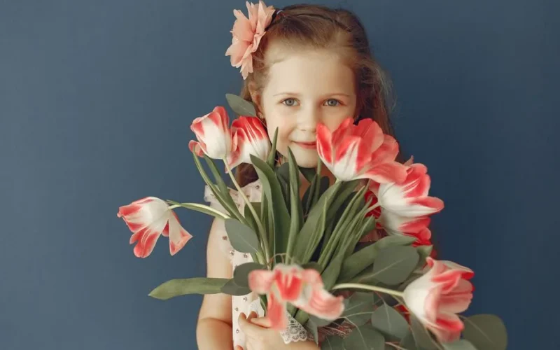 A young girl with a pink flower bouquet smiles warmly against a blue background.