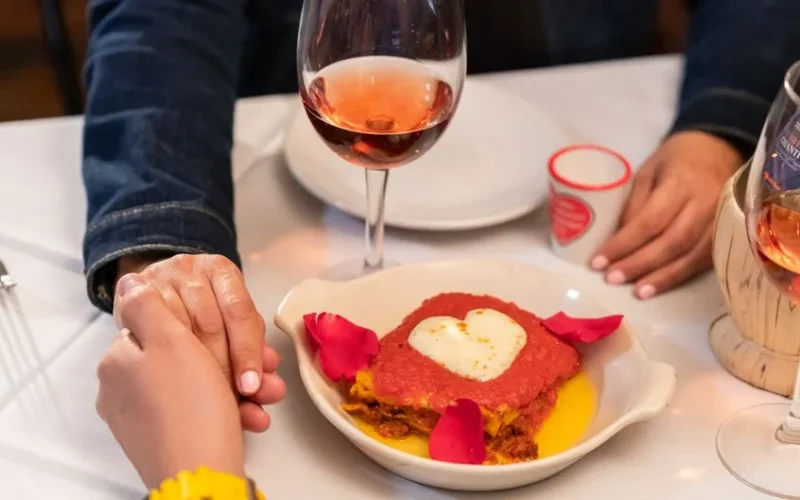 A romantic dinner scene with a heart-shaped dish, rose wine, and couple holding hands.